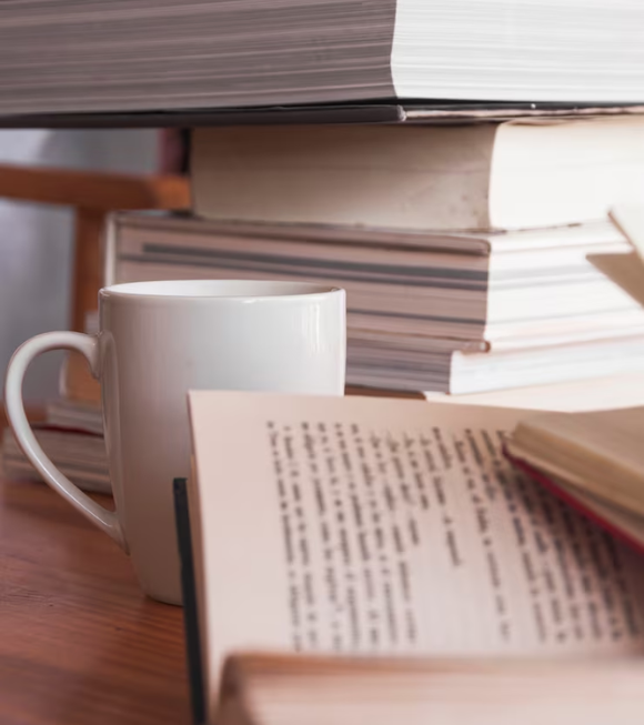 Stack of books on a wooden table, with bookshelves filled with more books in the background.